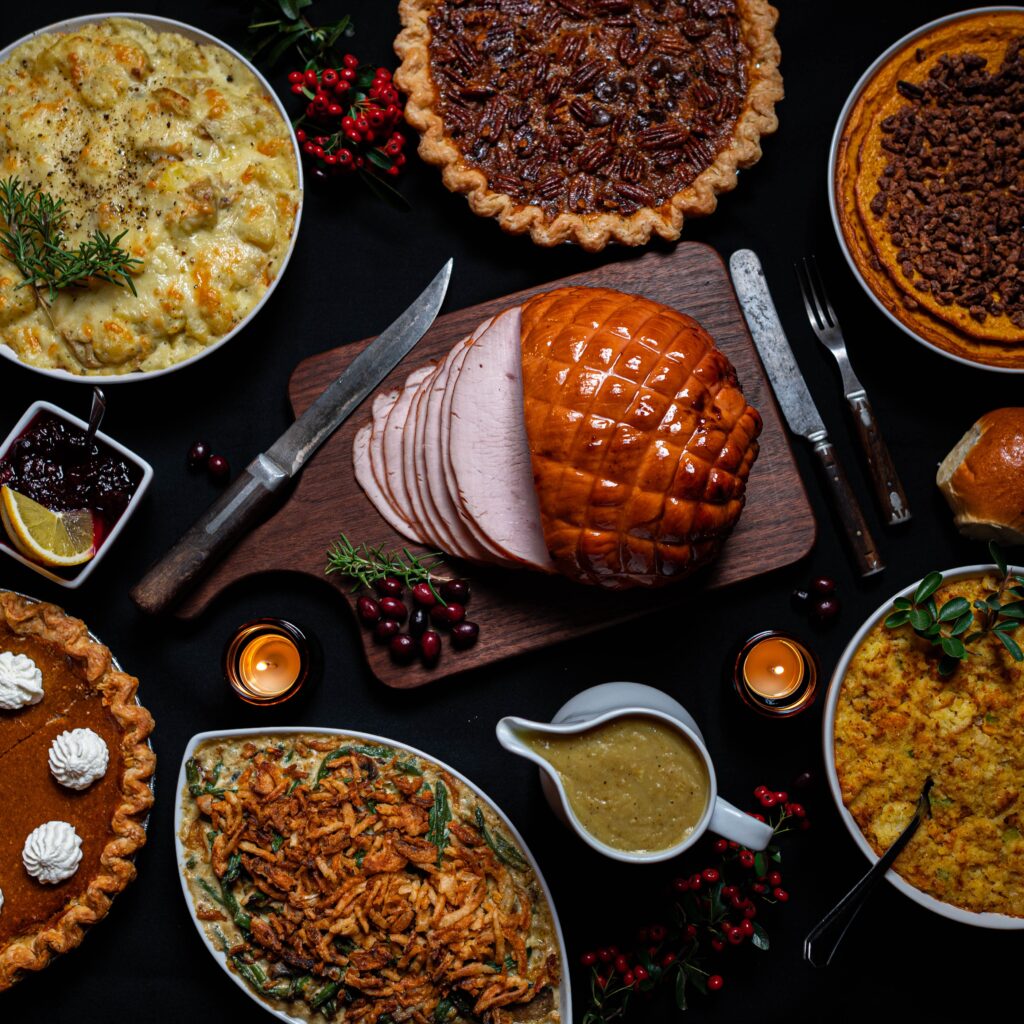 A ham sits in the middle of a table surrounded by other dishes like pie, casserole, gravy and fruits