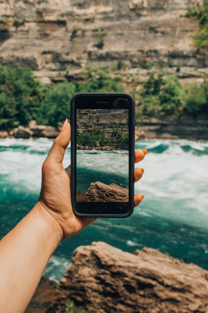 An iPhone in someone's hand where that person is taking a picture of sea or river and a rocky cliff