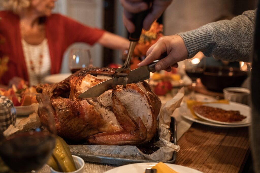 A turkey is being sliced by a person while another person in the background helps themselves to food