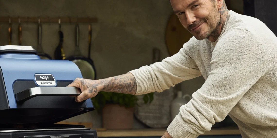 A-smiling-David-beckham-in-a-beige-sweater-bent-over-a-Ninja-Woodfire-outdoor-grill-placed-on-top-of-a-wood-top-table.