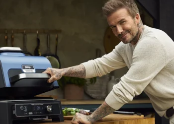 A-smiling-David-beckham-in-a-beige-sweater-bent-over-a-Ninja-Woodfire-outdoor-grill-placed-on-top-of-a-wood-top-table.