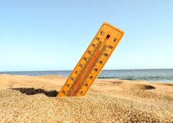 closeup-shot-of-yellow-wooden-thermometer-partly-buried-in-sand-with-blue-beach-backdrop