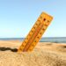 closeup-shot-of-yellow-wooden-thermometer-partly-buried-in-sand-with-blue-beach-backdrop